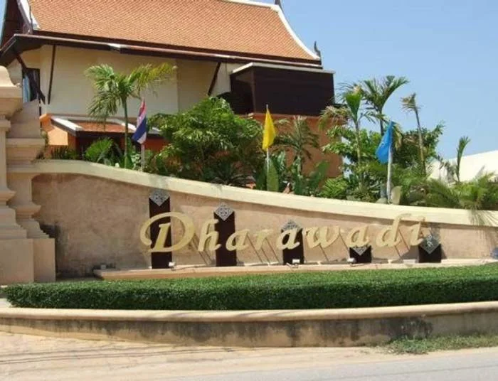 Dharawadi entrance sign with gold lettering on a stone wall, surrounded by green hedges and tropical plants, with a house in the background.